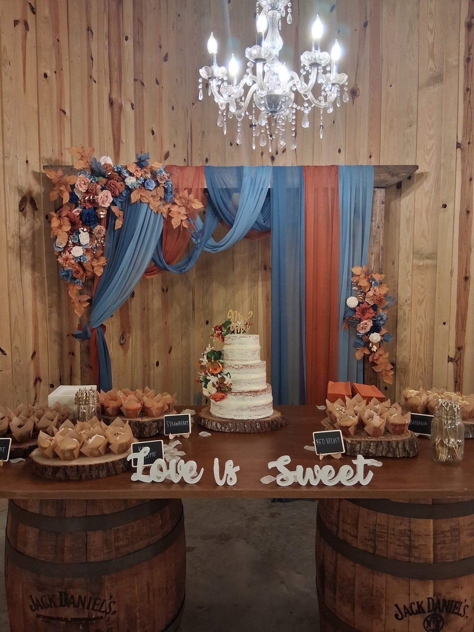 Rustic wedding dessert table with a three-tier cake on whiskey barrels and a Love is Sweet sign