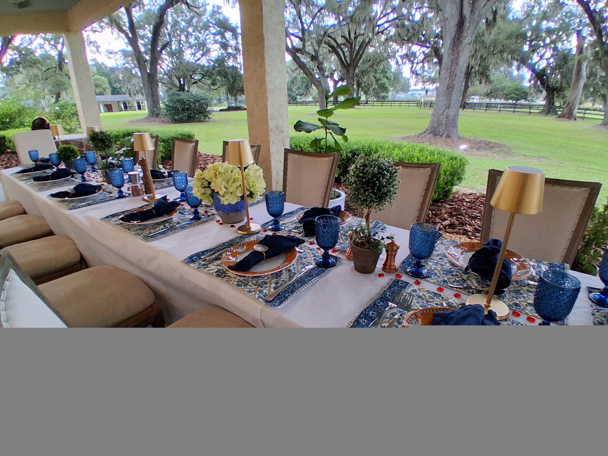 Long outdoor patio dinner table with blue glassware and gold lamps overlooking a lawn